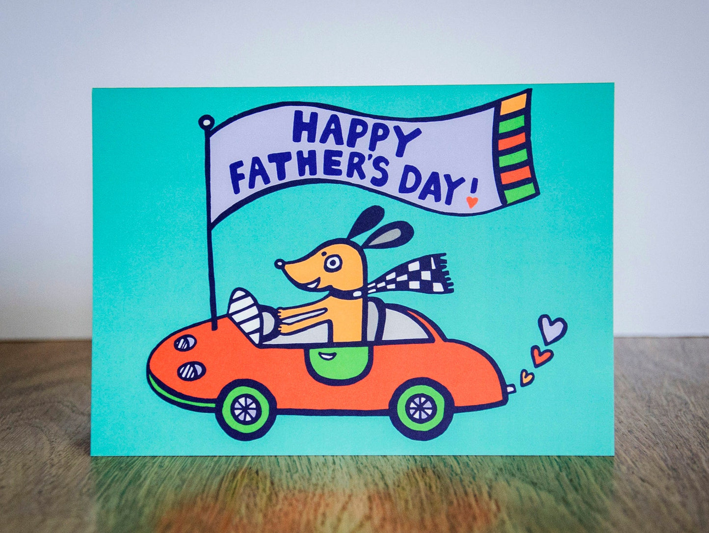 A turquoise card featuring a dog driving a red sports car with a flag that reads "Happy Father's Day!"