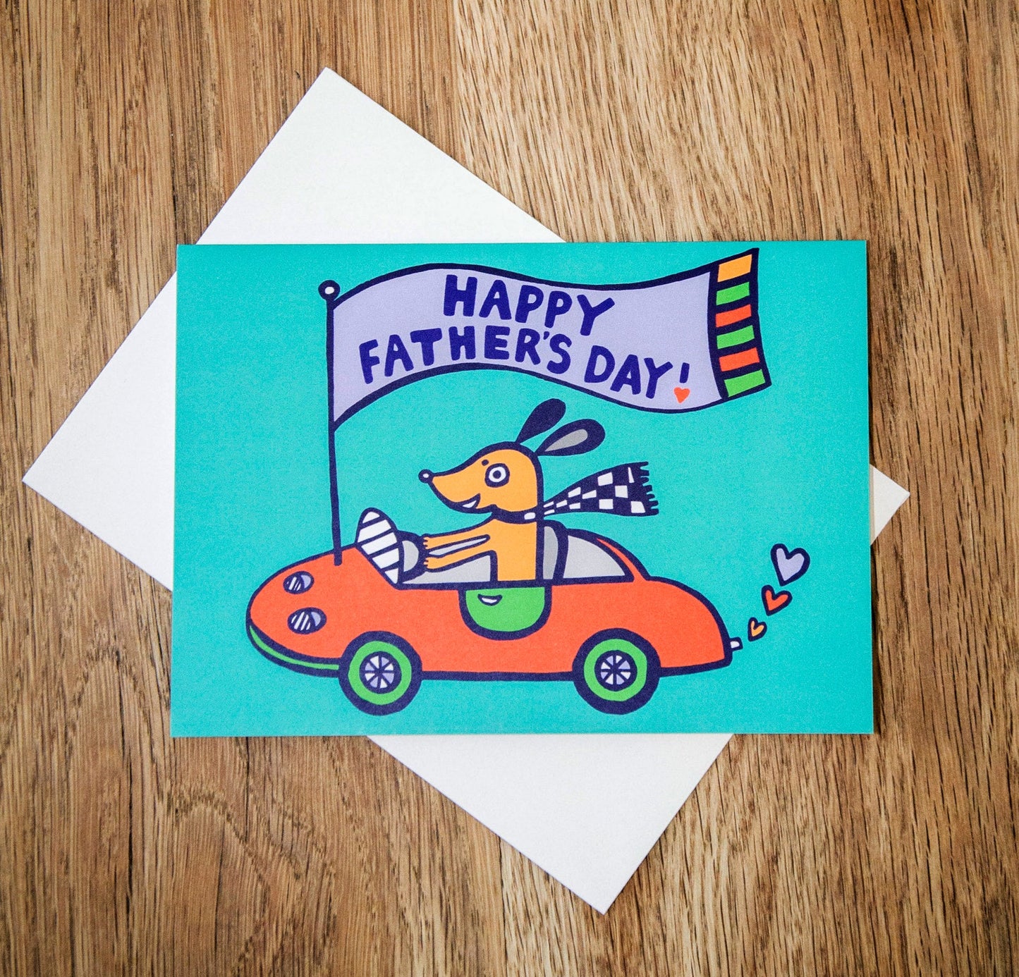 A turquoise card featuring a dog driving a red sports car with a flag that reads "Happy Father's Day!"