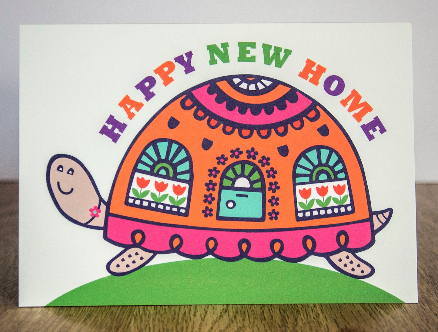A card depicting a colourful happy tortoise with a door and windows on its brightly decorated shell. Curved above the shell reads the text "Happy New Home".