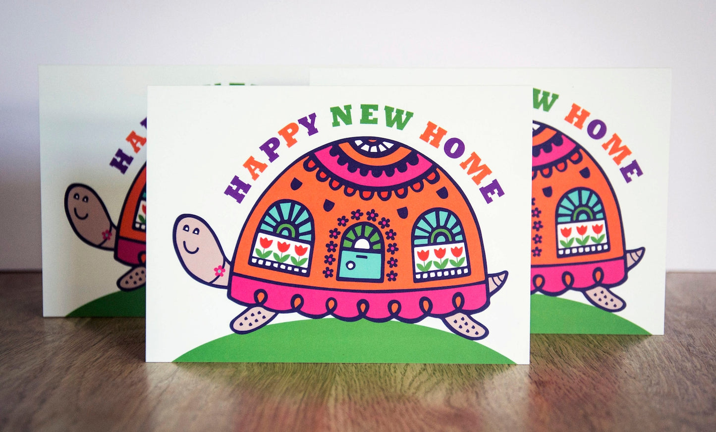 A card depicting a colourful happy tortoise with a door and windows on its brightly decorated shell. Curved above the shell reads the text "Happy New Home".