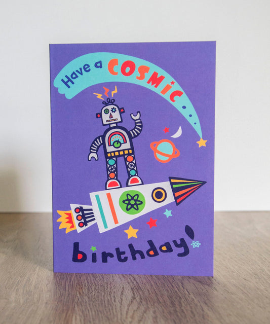 A vibrant card that depicts a robot standing atop a rocket in space. Above the robot there is a shooting star. The text on the card reads "Have a cosmic birthday!"