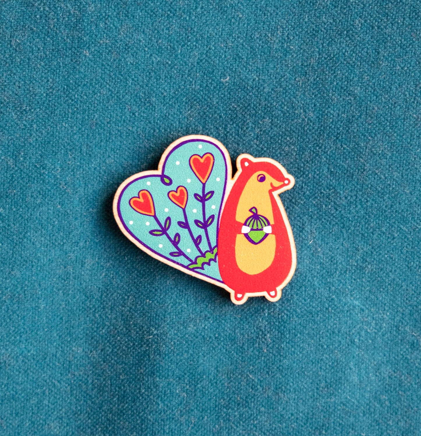 A brightly panted art deco inspired wooden brooch of a red squirrel with blue tail, smiling and holding an acorn.