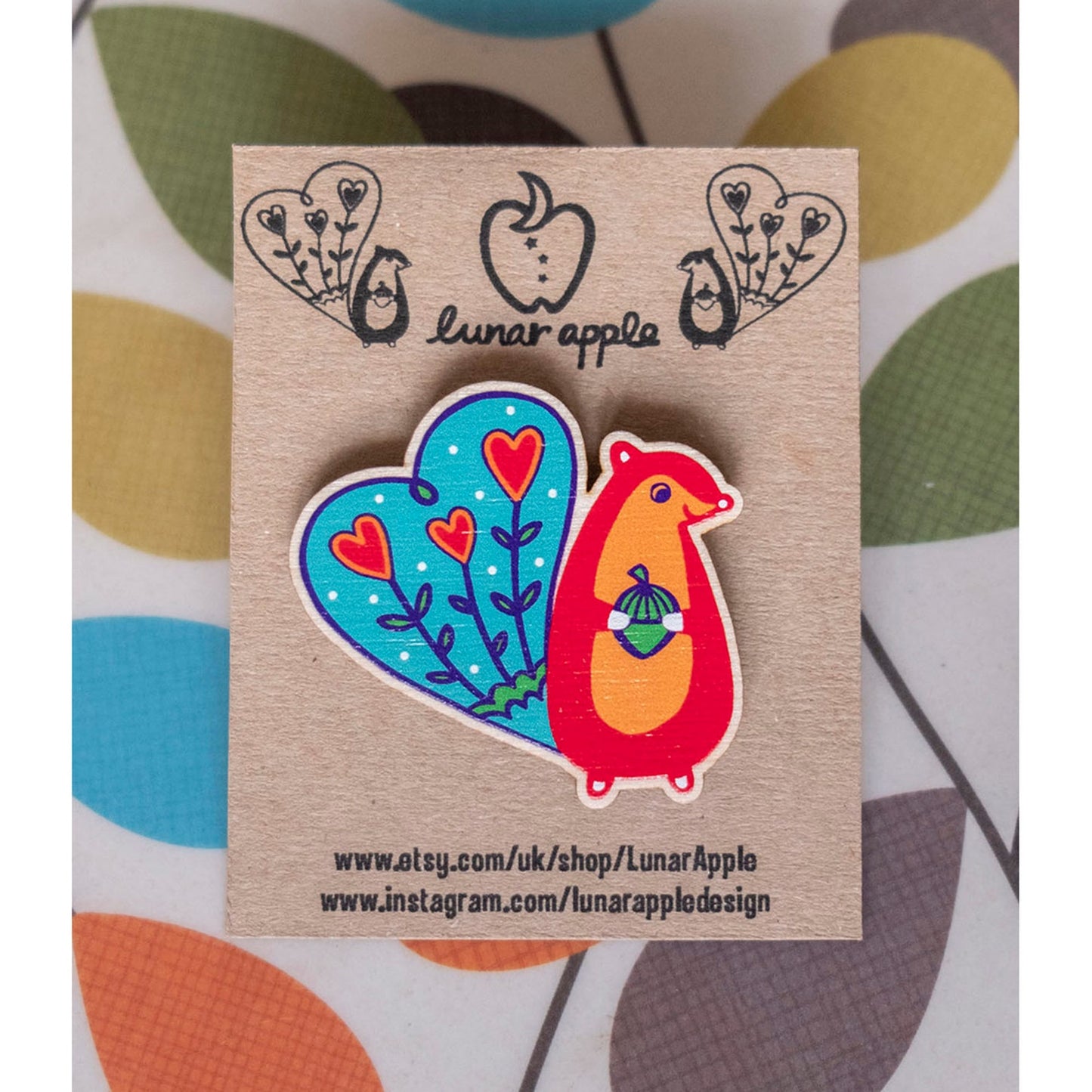 A brightly panted art deco inspired wooden brooch of a red squirrel with blue tail, smiling and holding an acorn. The brooch is pinned to a brown card backing.