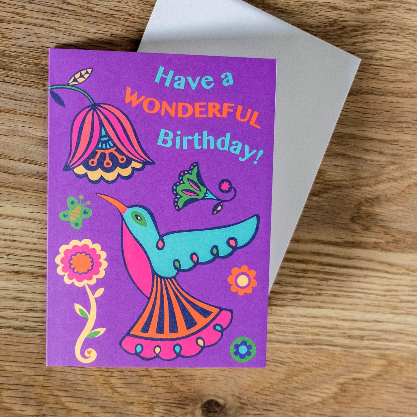 A purple card with a blue, pink and orange art deco style hummingbird looking up at a flower. Above reads the text "Have a Wonderful Birthday!"