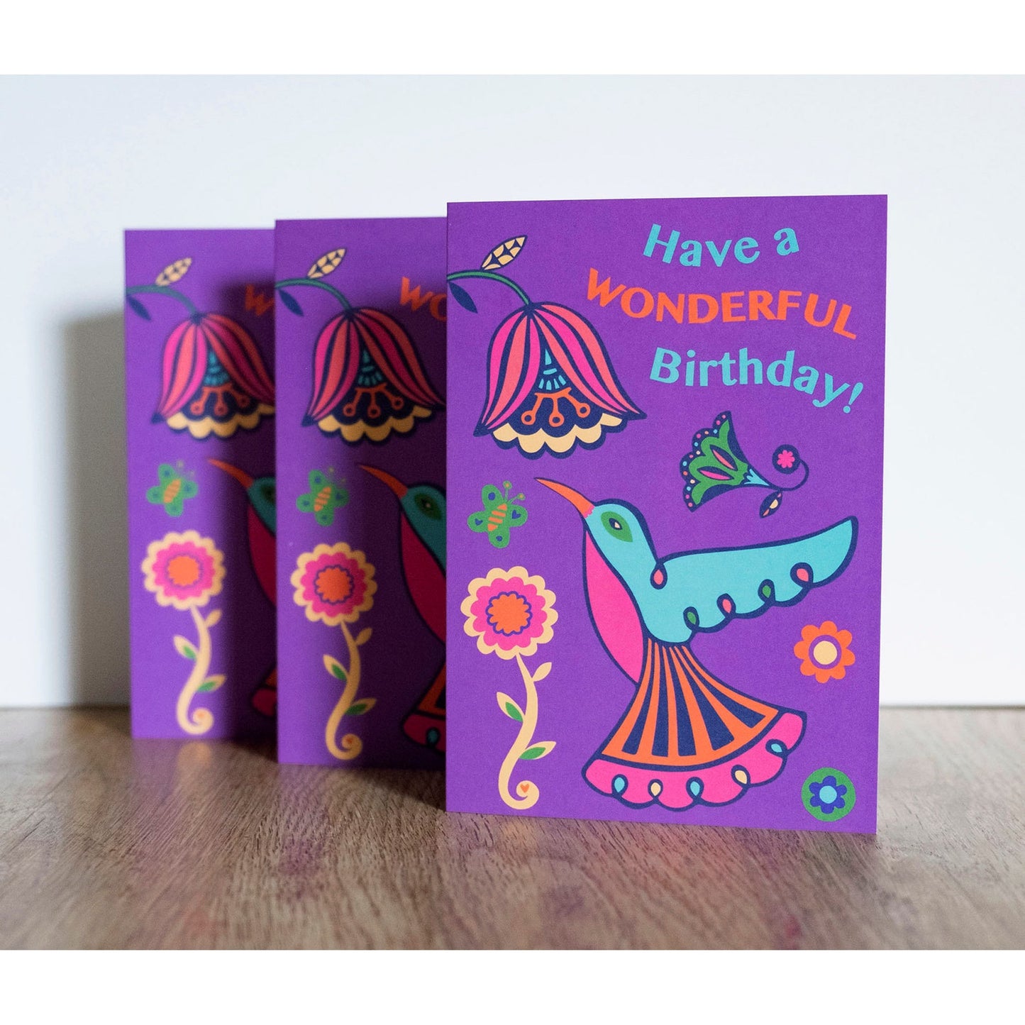 A purple card with a blue, pink and orange art deco style hummingbird looking up at a flower. Above reads the text "Have a Wonderful Birthday!"