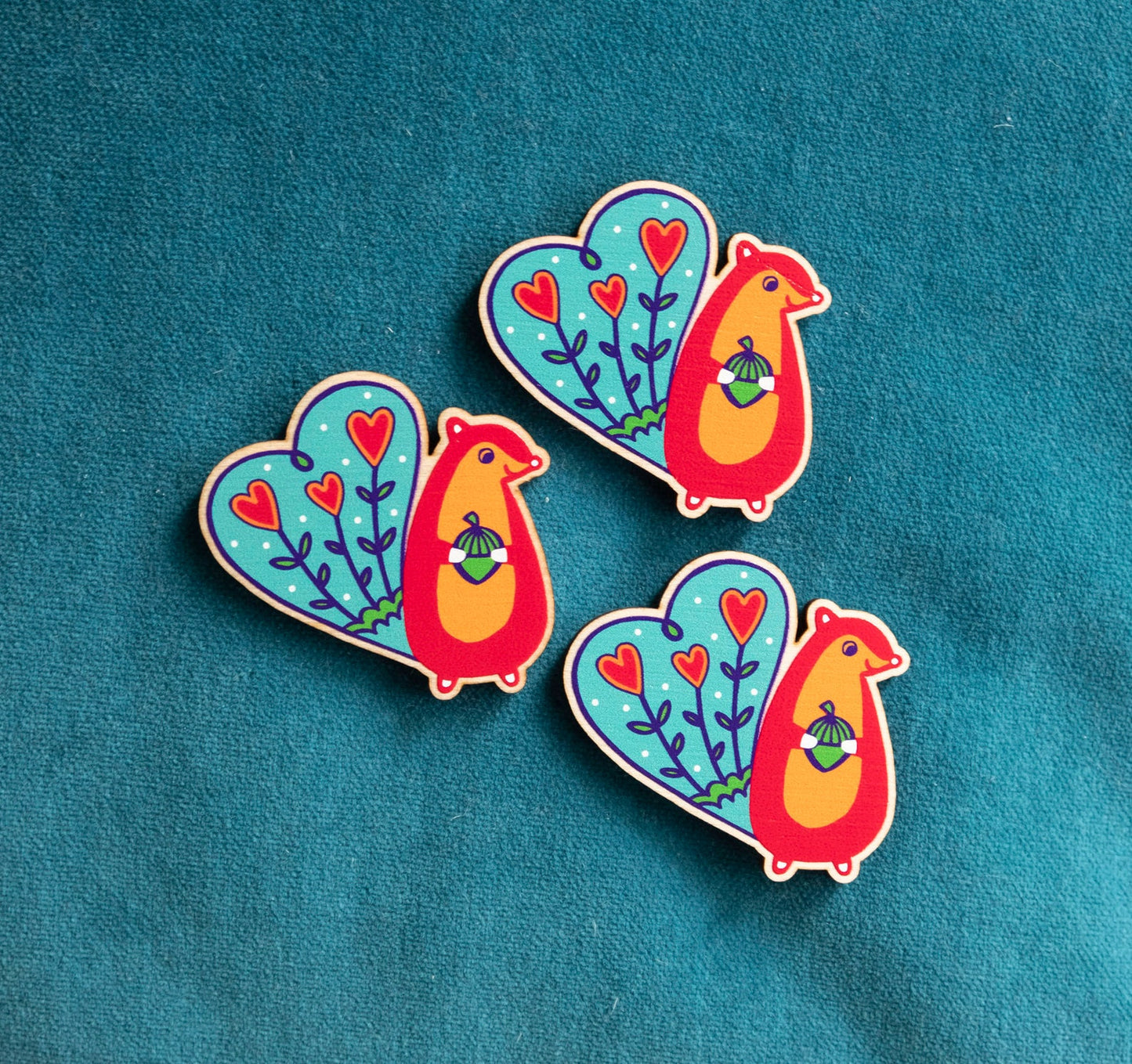 Three brightly panted art deco inspired wooden brooches of a red squirrel with blue tail, smiling and holding an acorn.