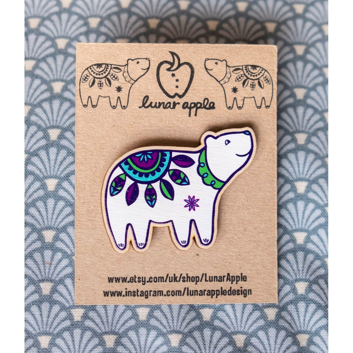 A brightly painted wooden brooch of an art deco inspired smiling polar bear pinned to a brown card backing.
