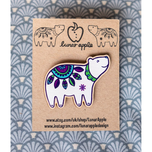 A brightly painted wooden brooch of an art deco inspired smiling polar bear pinned to a brown card backing.