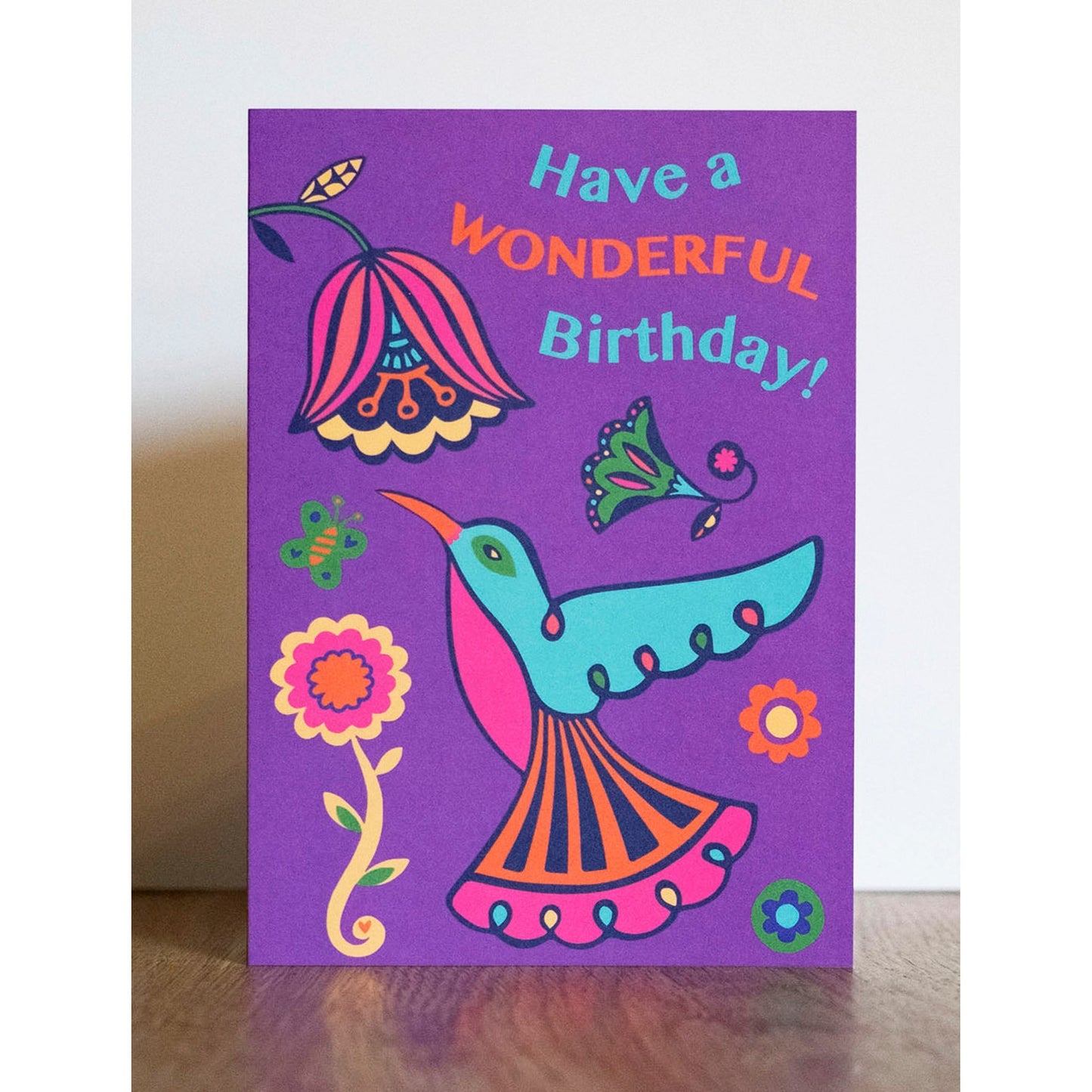 A purple card with a blue, pink and orange art deco style hummingbird looking up at a flower. Above reads the text "Have a Wonderful Birthday!"