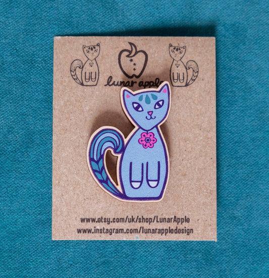 A brightly painted wooden brooch of an art deco inspired grey cat pinned to a brown card backing.
