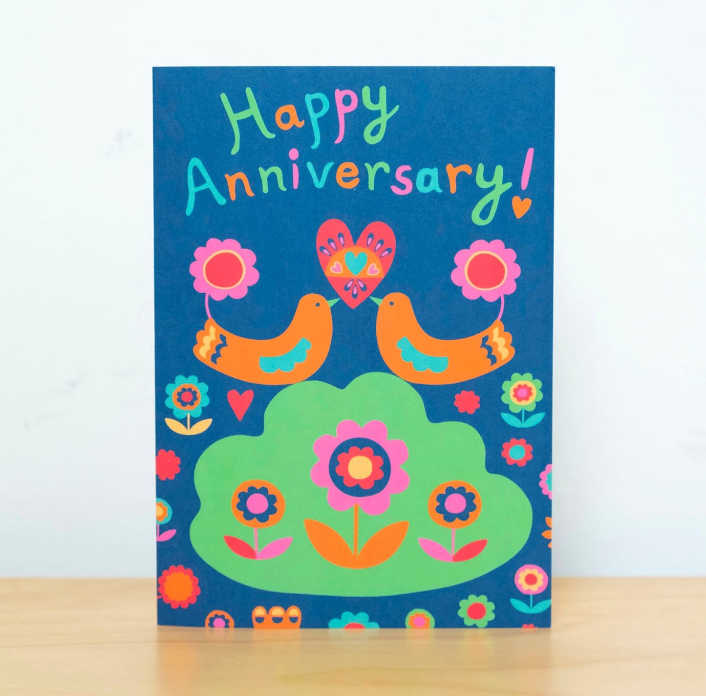 A card depicting two orange art deco birds holding up a heart together with their beaks. Above them reads the text "Happy Anniversary!"
