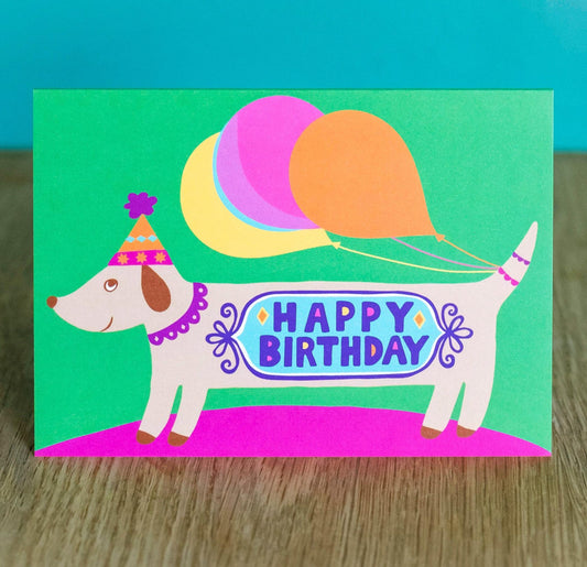 A card depicting a smiling sausage dog with balloons attached to its tail, with the message "happy birthday" across the body.