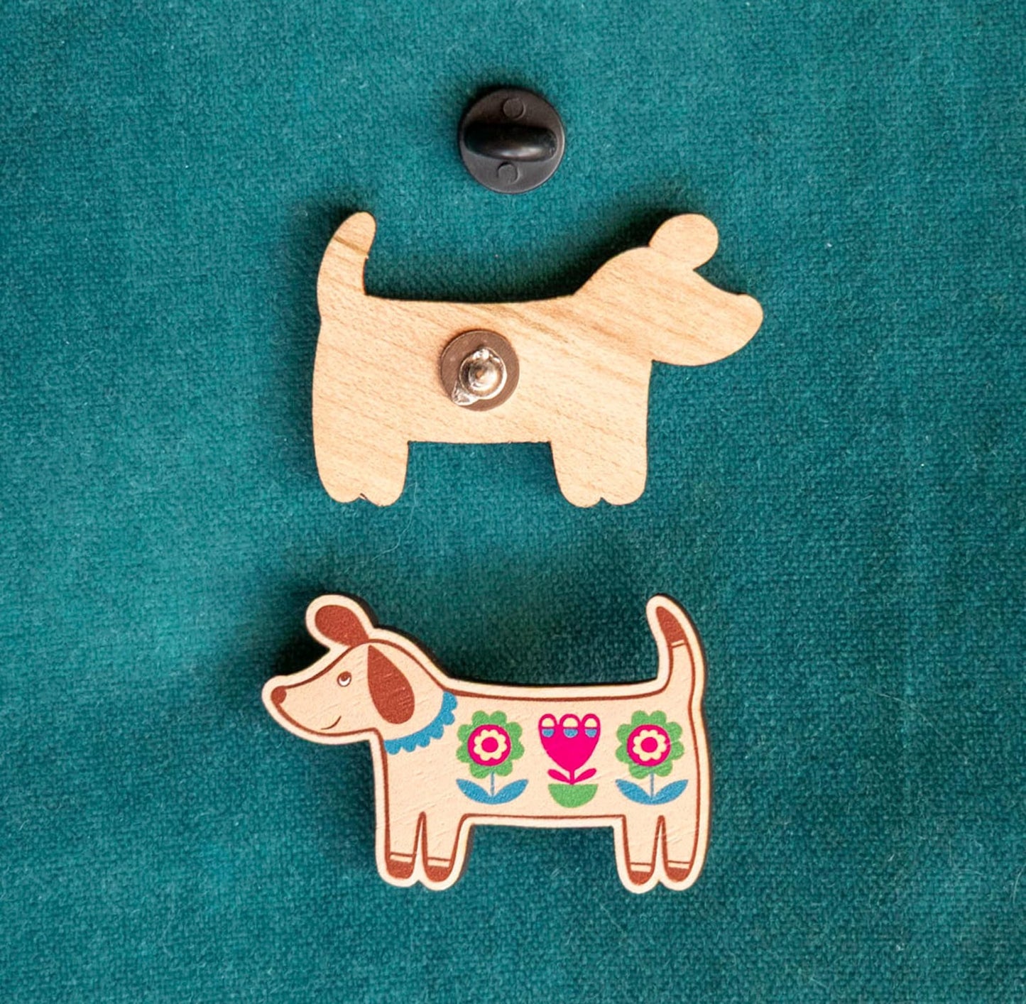 The front and back of a wooden dog-shaped pin against a teal background, with a rubber fastening.