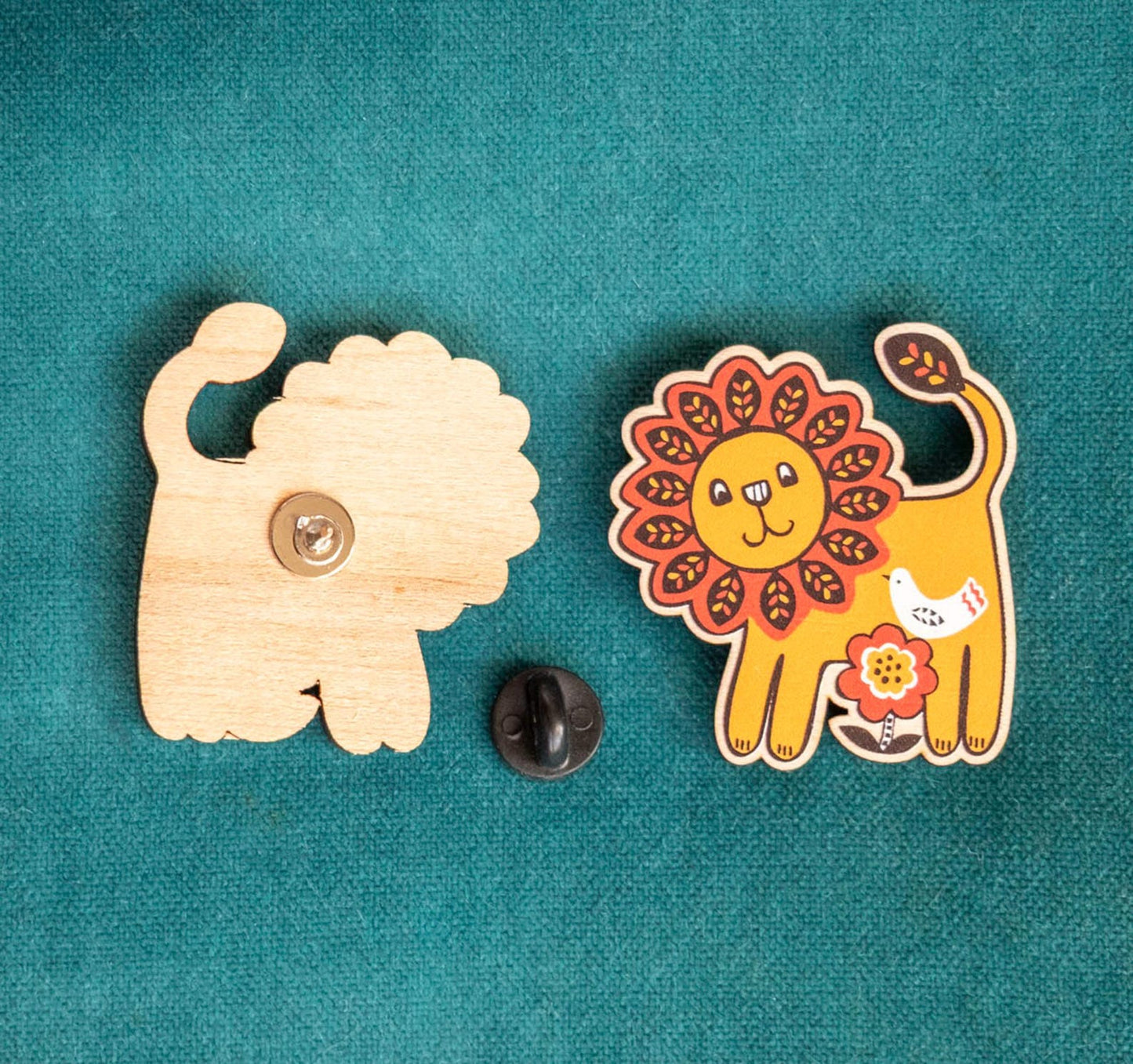 The front and back of a colorful, smiling lion-shaped wooden pin with rubber fastening, placed against a teal background.