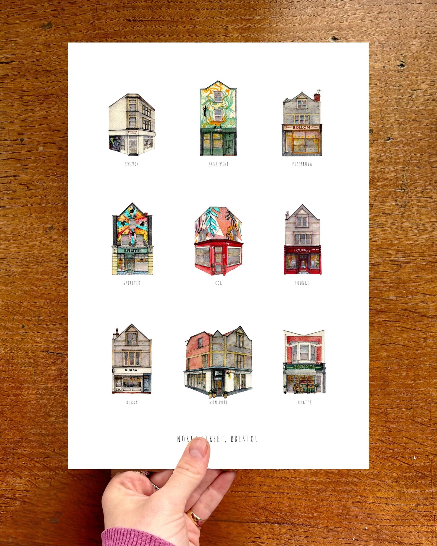 Poster of illustrated buildings held by a hand on a wooden surface