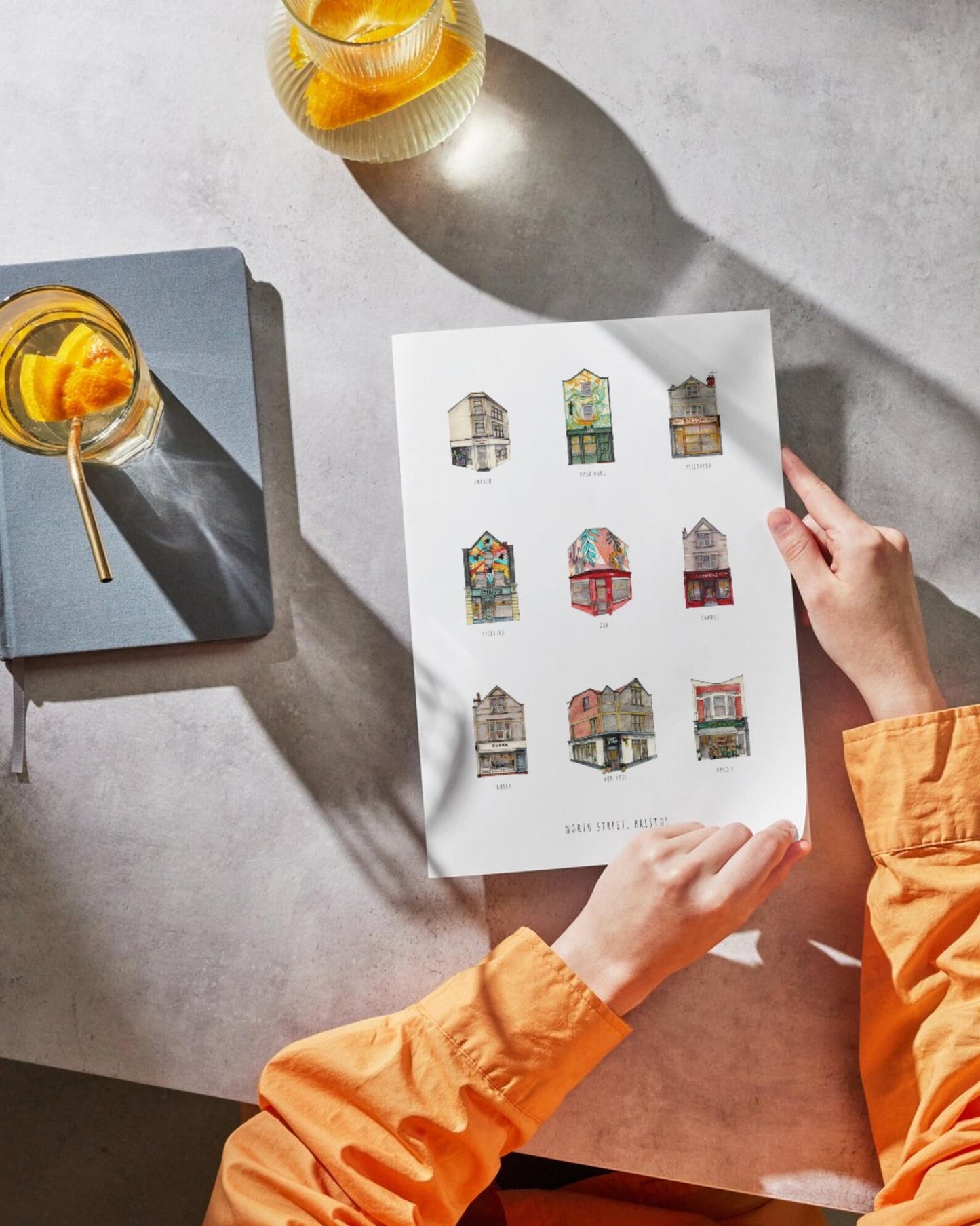 Person holding a sheet of illustrated house prints on a textured surface with glasses of yellow liquid.