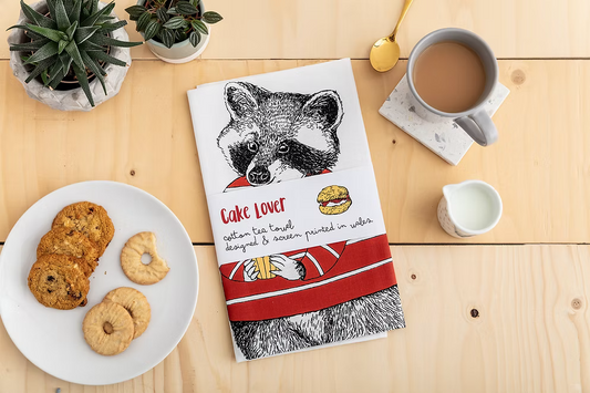 Tea towel with raccoon design on a wooden table with cookies and coffee.