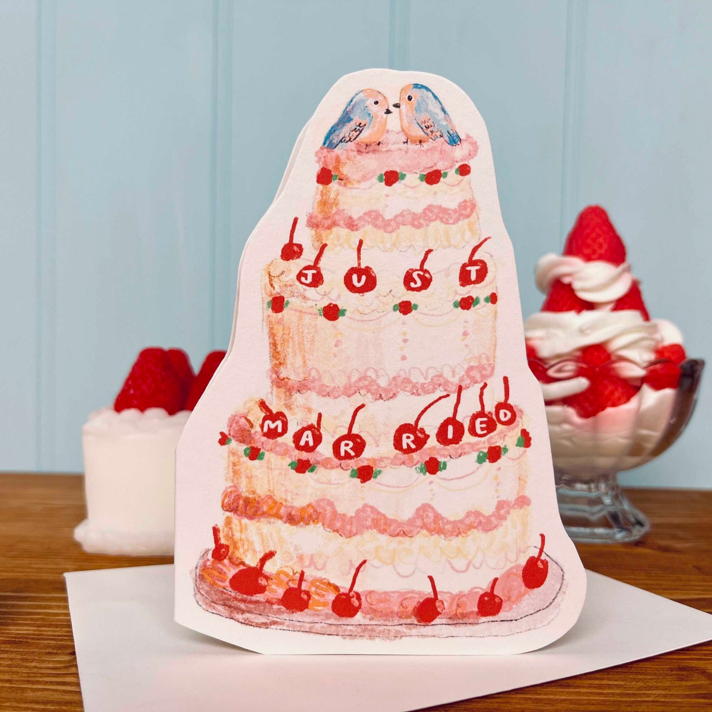 Card shaped like a wedding cake with cherries and 'MARRIED' text on a wooden surface.