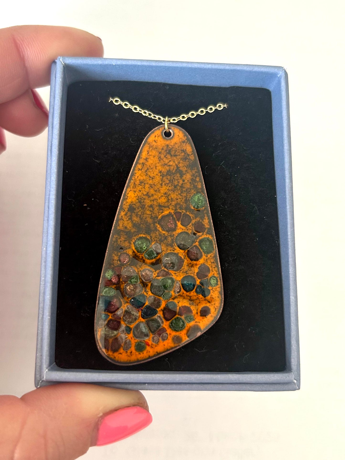 A pebble-shaped orange enamel pendant with glass chips fused into it, resting in a jewellery box.