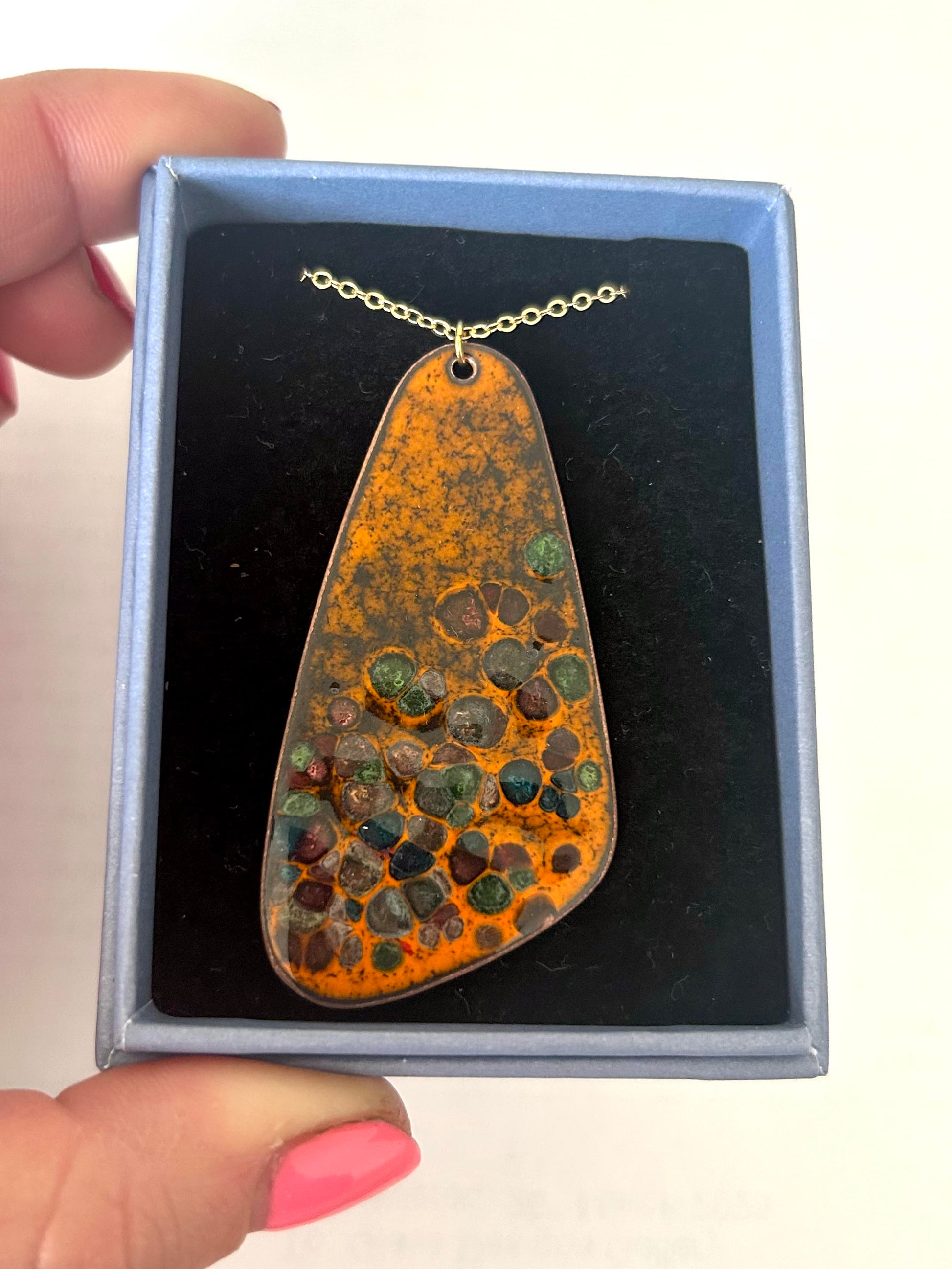 A pebble-shaped orange enamel pendant with glass chips fused into it, resting in a jewellery box.