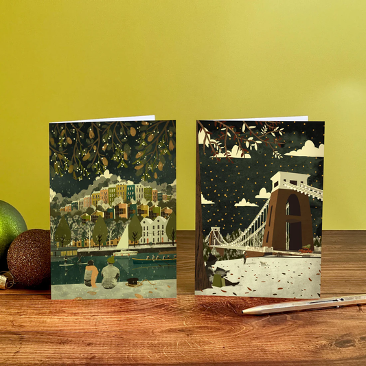 Two greeting cards with artistic designs of a cityscape and a bridge on a wooden surface with a yellow wall background.