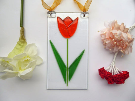 A handcrafted glass hanging decoration featuring a tulip flower design, with a copper hook and colored ribbon, set against a leafy backdrop.