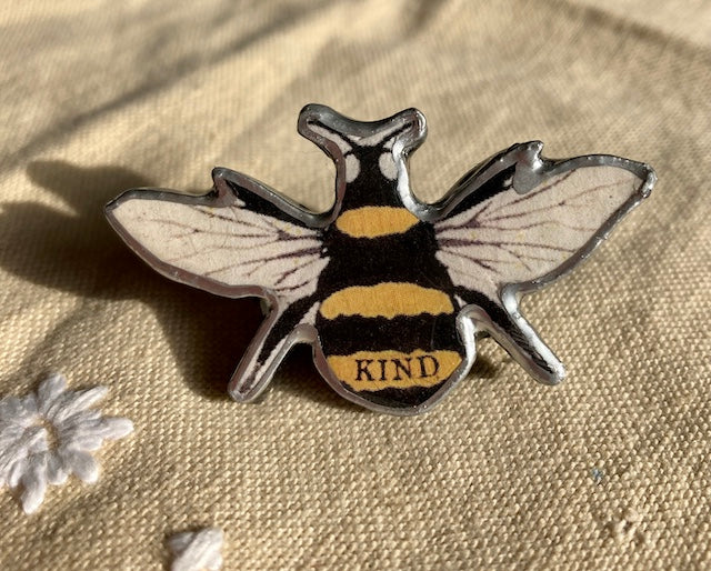 Bumble bee brooch with the text Kind on its body. Handmade in Cardiff by Ellymental. Layered paper, metal and resin