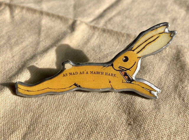 Brooch made from layered paper and steel. Laquered with a thick resin. A yellow hare in full scamper with the text "as mad as a march hare' on its body.