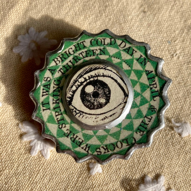 This George Orwell Eye brooch is handmade by Elemental a Cardiff artist. Made from layered paper and metal a laquered with resin to seal. The text around the eye reads It was a bright cold day in April and the clocks were striking the thirteen.