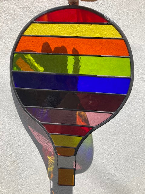 A large stained glass hot air balloon with rainbow stripes held against a brick wall background.