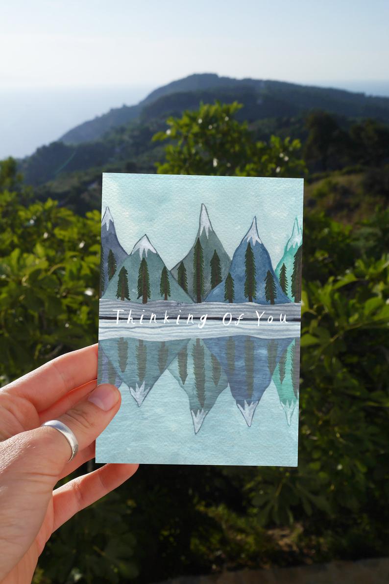 watercolour greetings card with mountains and trees reflected in a lake and the words ''thinking of you''