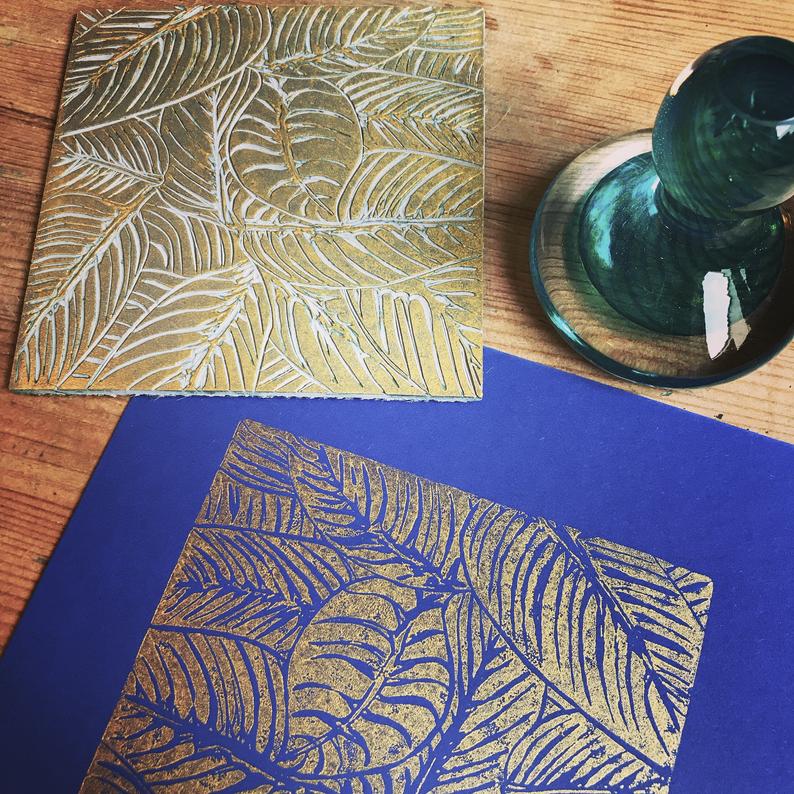 Trelawney Designs hand cut lino print with water based ink. Botanical leaves in Gold on a Dark blue back ground. Framed in a white frame. Edit alt text