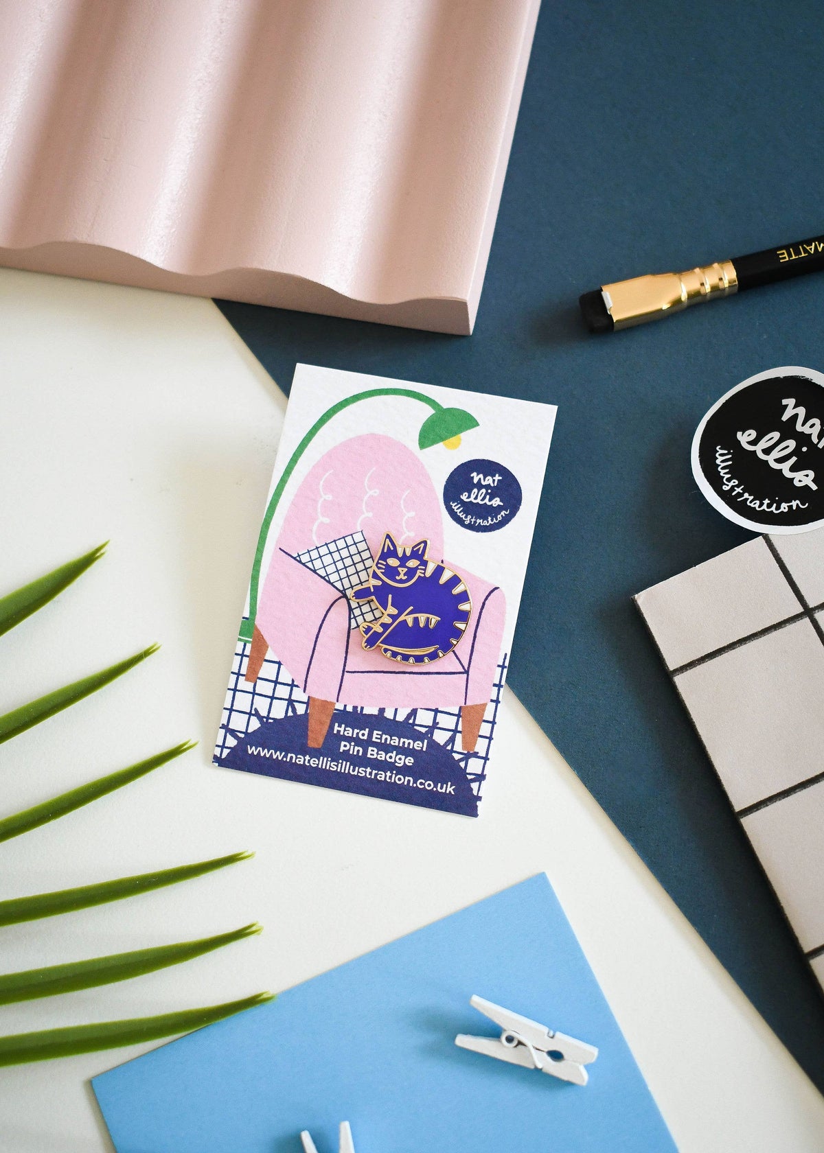 A flat-lay product photo of a hand enamel pin badge on a white card, styled with a pink sheet, blue paper, a pencil, and a sticker on a dark blue surface.