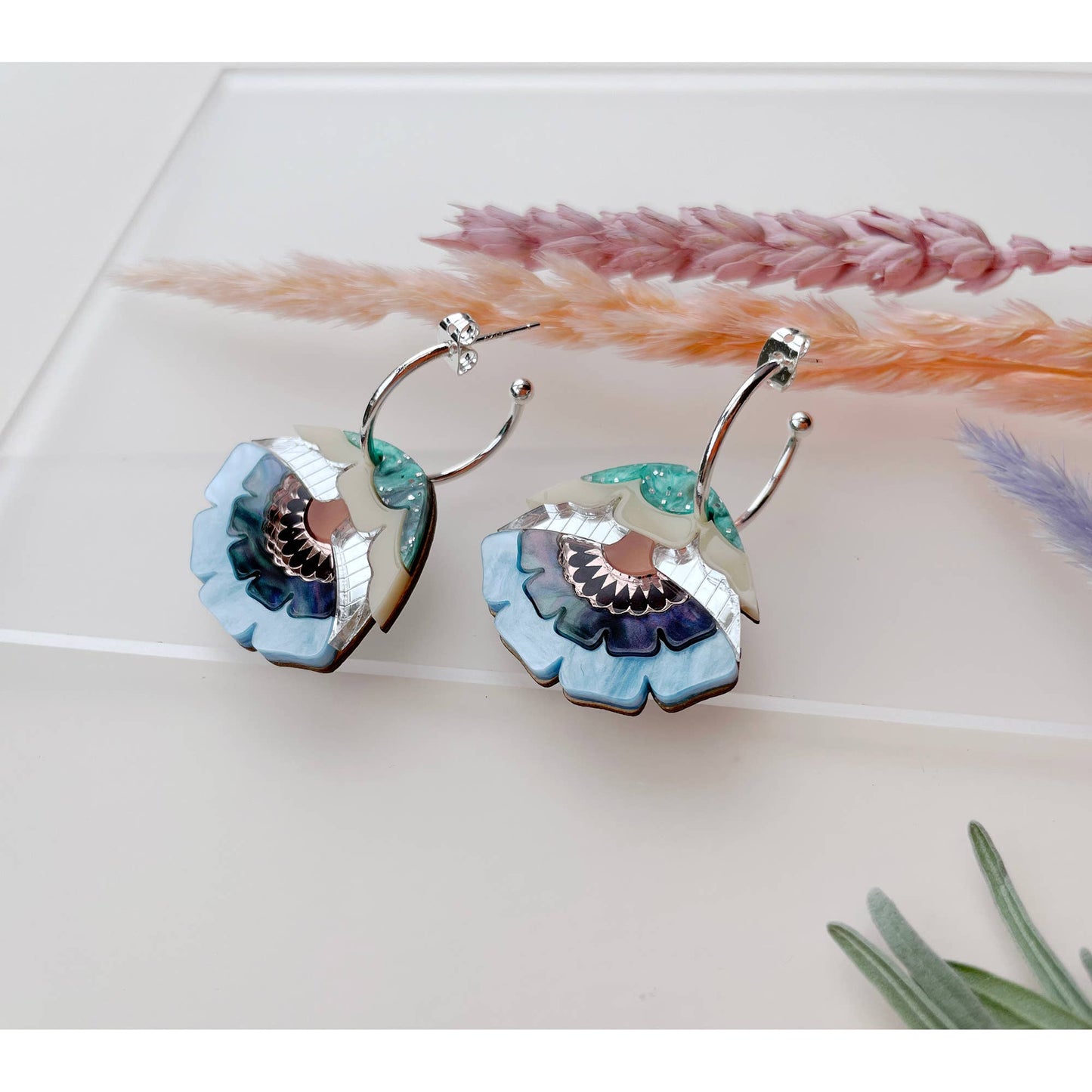 A pair of silver hoop earrings with layered flower-shaped drops in light blue, teal, purple, and cream, displayed on a white surface with dried grass stems.