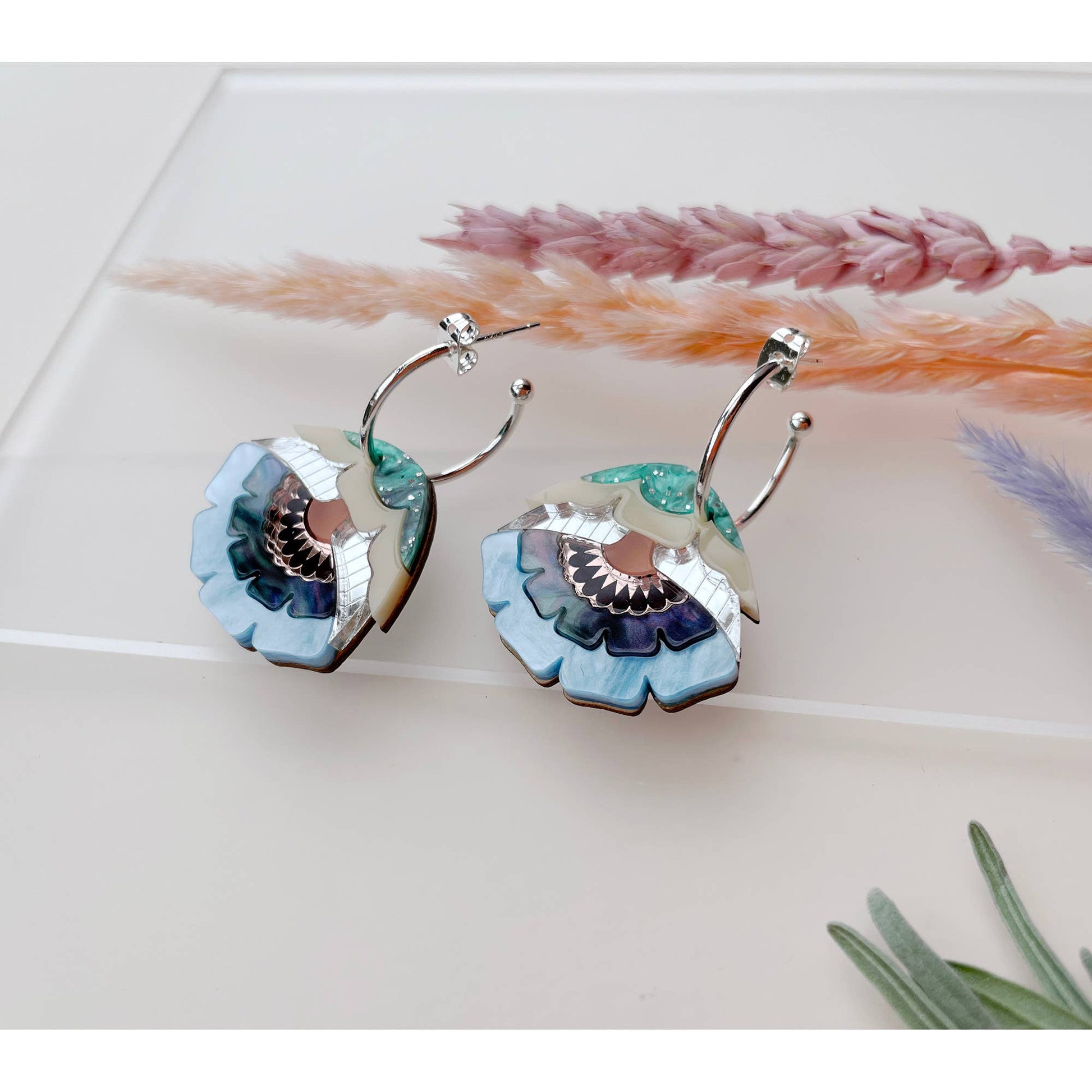 A pair of silver hoop earrings with layered flower-shaped drops in light blue, teal, purple, and cream, displayed on a white surface with dried grass stems.