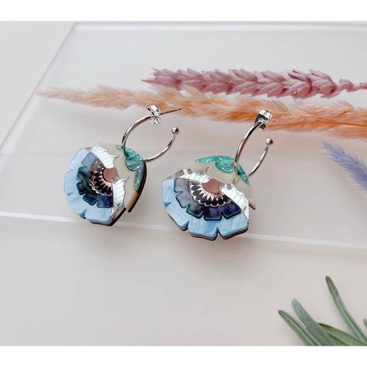 A pair of silver hoop earrings with layered flower-shaped drops in light blue, teal, purple, and cream, displayed on a white surface with dried grass stems.
