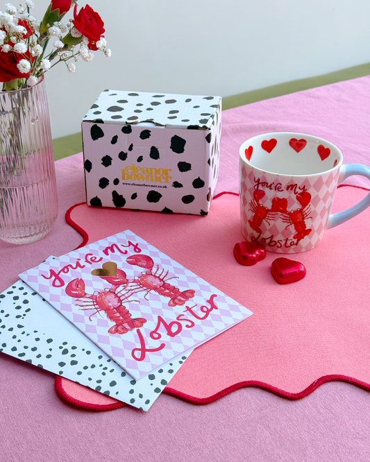 Valentine's Day-themed mug, card, and box on a pink surface with red flowers.
