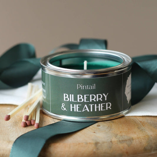 Candle in a green tin labeled 'Bilberry & Heather' with matches and ribbons on a wooden surface.
