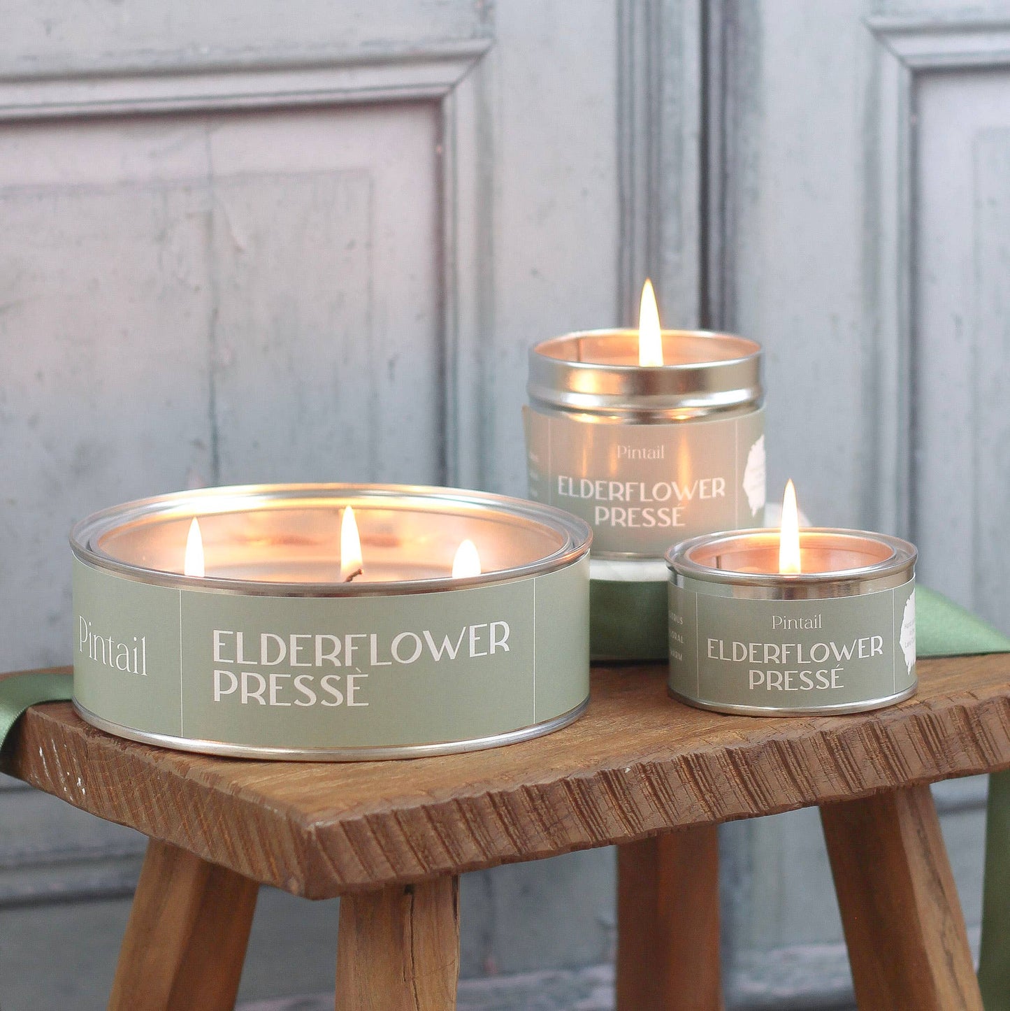Three lit candles in tin containers labeled 'Elderflower Presse' on a wooden stool.