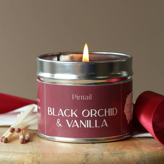 Candle in a tin labeled 'Black Orchid & Vanilla' by Pintail, with matches and red ribbon on a wooden surface.