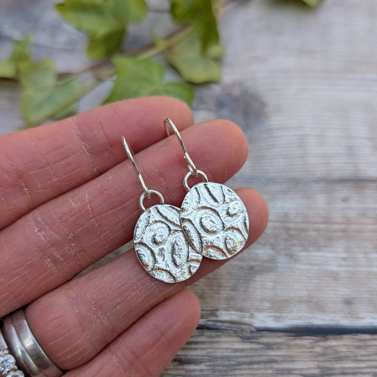 A pair of silver drop earrings with textured circular pendants, held in a person's hand against a rustic wooden background with blurred green leaves.