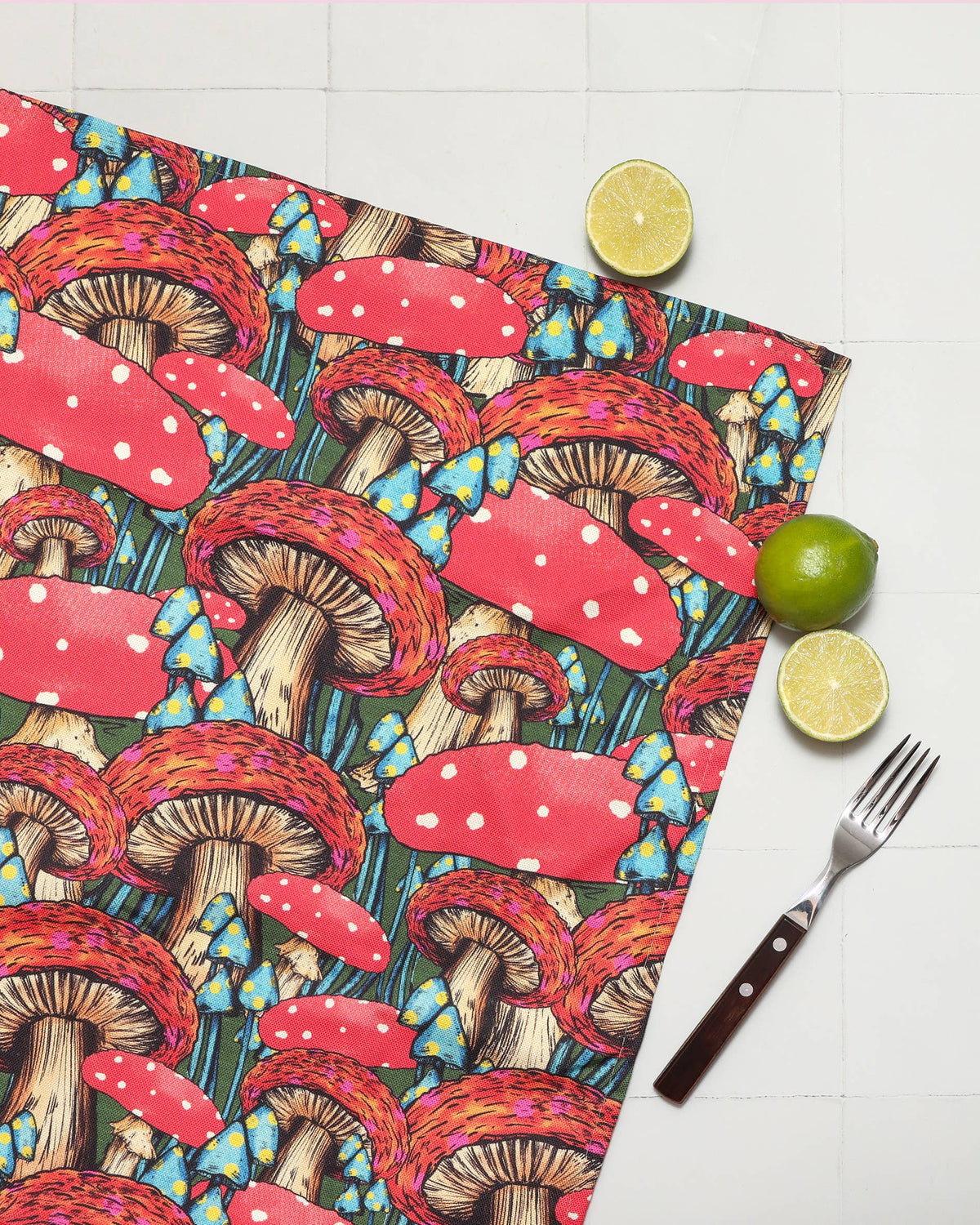 A square placemat with a whimsical mushroom print, styled on a white tiled surface with a fork, a lime, and a lemon half.