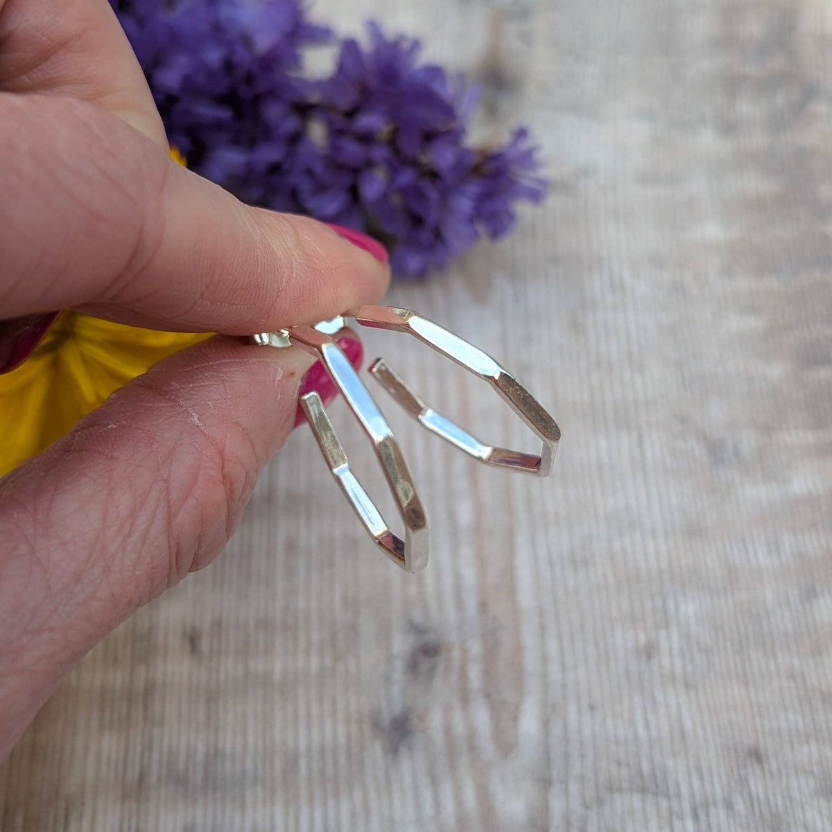A hand holding a pair of silver-toned geometric hoop earrings against a light wood background with blurred purple flowers.