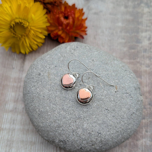 A pair of silver dangle earrings with small polished rose-gold heart charms, displayed on a smooth gray stone with yellow and orange flowers in the background.