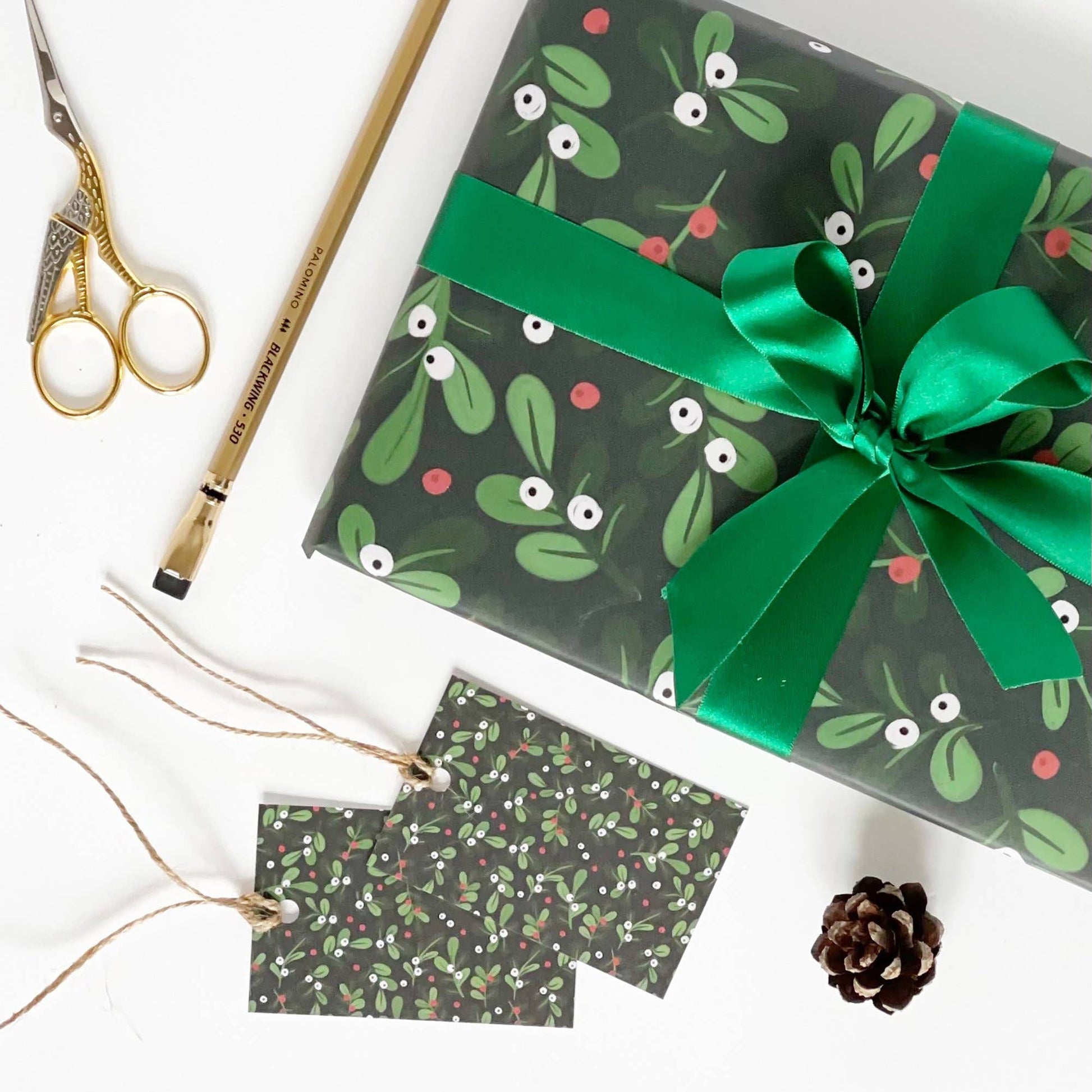 A wrapped gift box with a green ribbon and bow, shown in a styled flat lay with matching patterned gift tags, gold scissors, a pencil, and a small pinecone on a white background.
