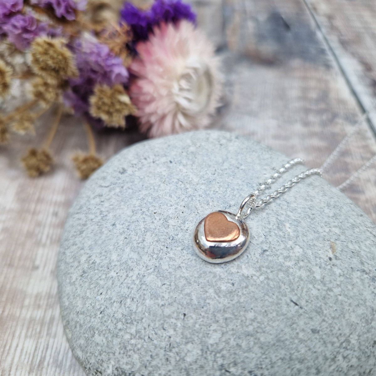 A silver necklace with a polished oval pendant featuring a small rose gold heart in the center, resting on a smooth gray stone with dried flowers in the background.