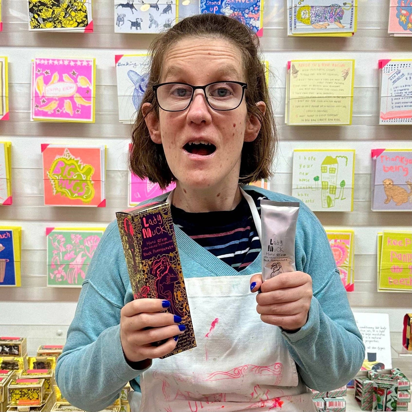 Person holding Lady Muck products in a store with colorful greeting cards on the wall.