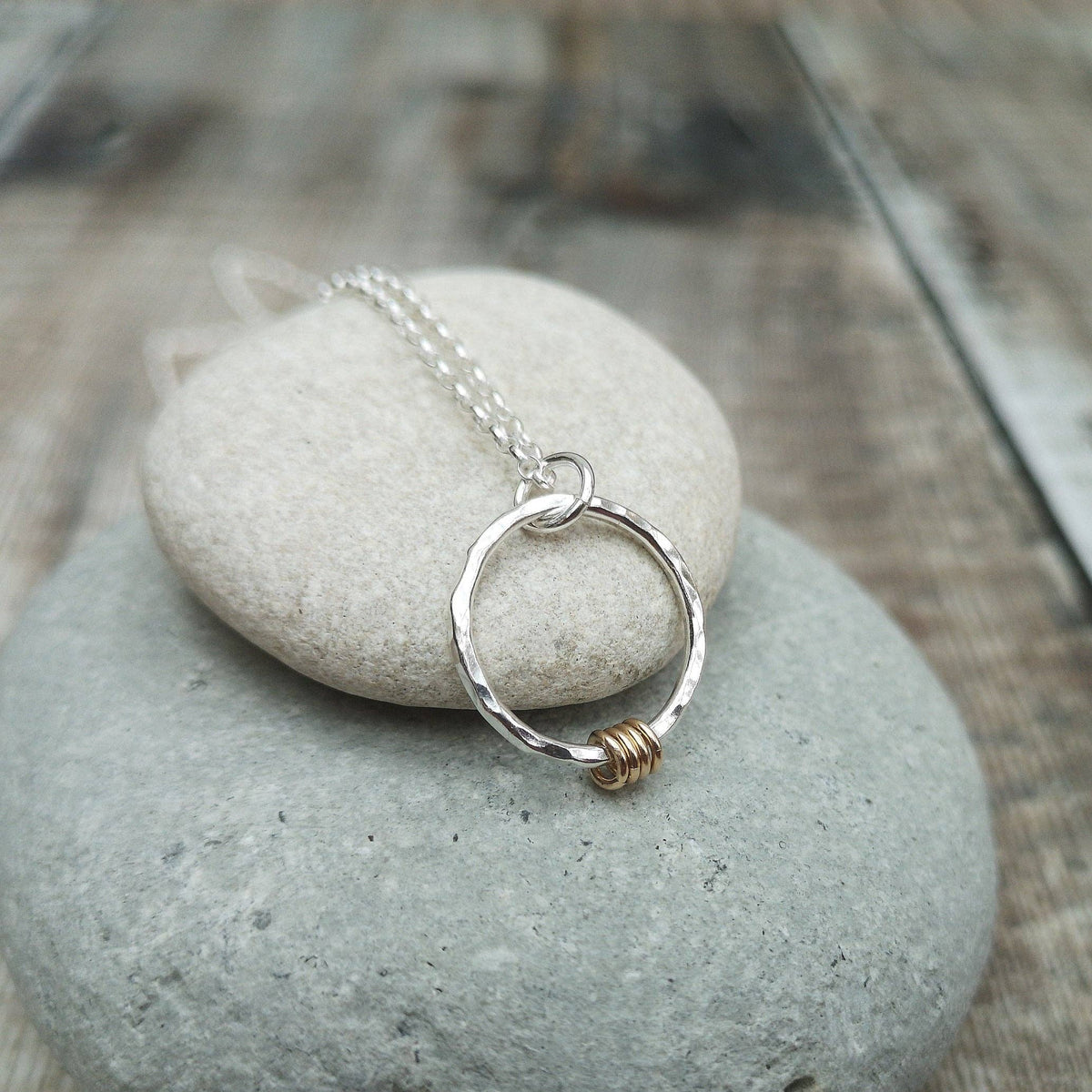 A silver necklace with a hammered circular pendant and a small gold wire accent, displayed on smooth gray stones against a blurred rustic wooden background.
