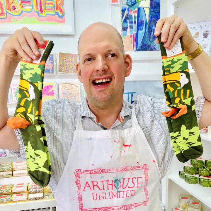 A smiling man wearing a white apron with 'ARTHOUSE UNLIMITED' stands in a craft or sock workshop, holding up a pair of green camouflage patterned socks with bright orange and yellow accents.