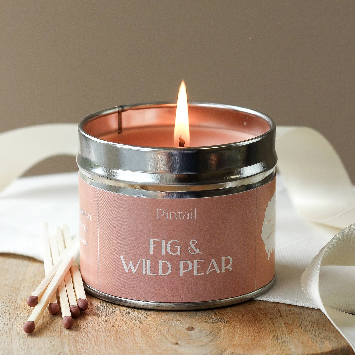 Candle in a tin labeled 'Fig & Wild Pear' with matches on a wooden surface.